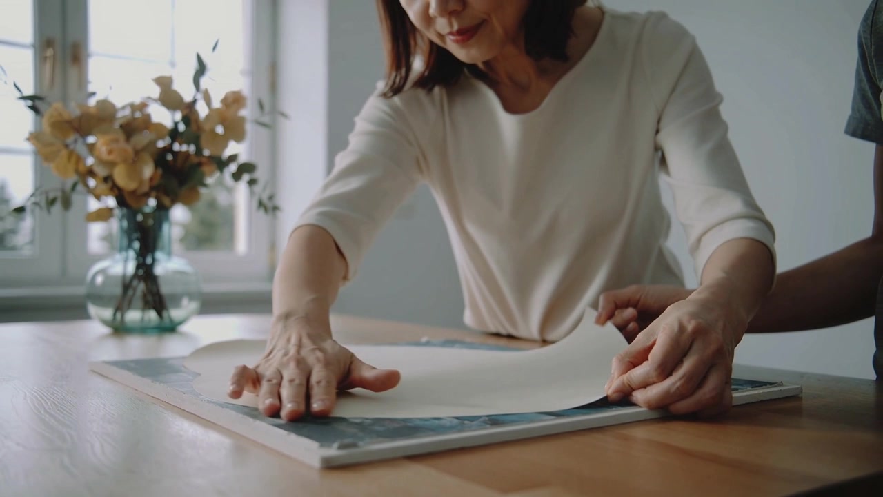 Smoothing woman wearing cream top aligning paper on frame by window, assistant holding right edge