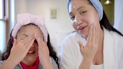 happy biracial mother and daughter with headbands putting cream on faces in sunny bathroom