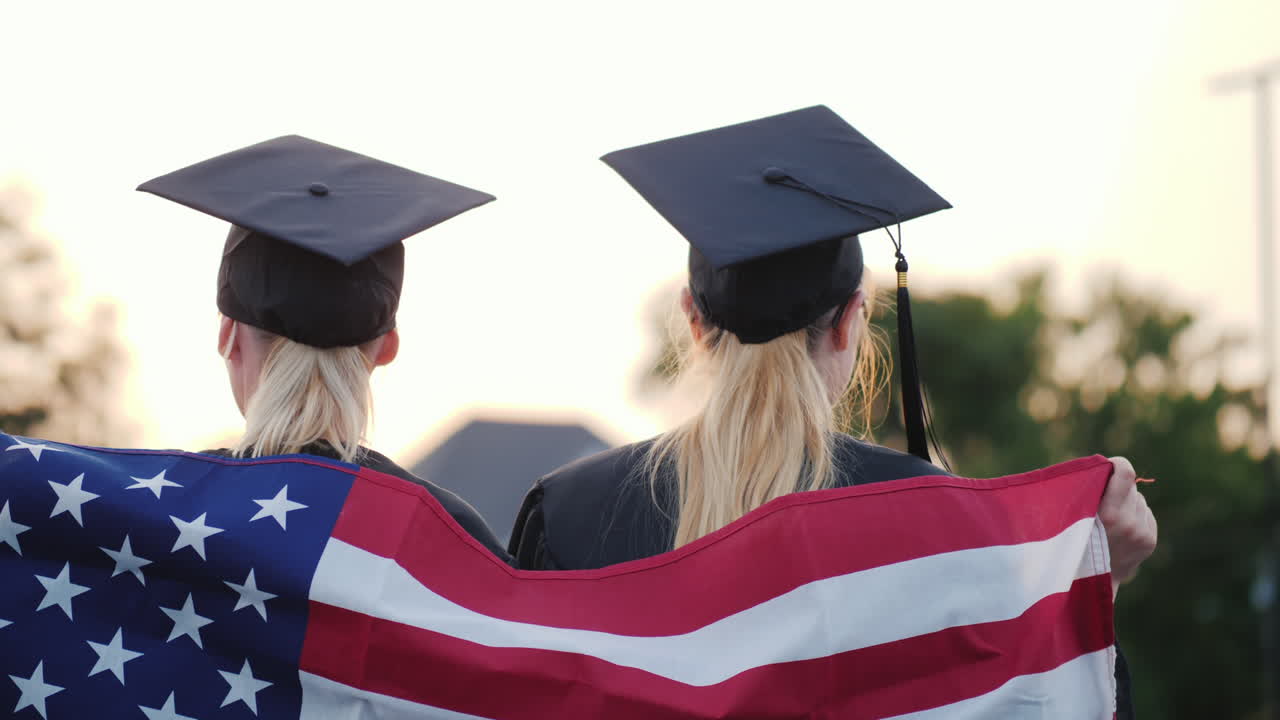 dos mujeres graduadas universitarias con la bandera de estados unidos en los hombros vista trasera