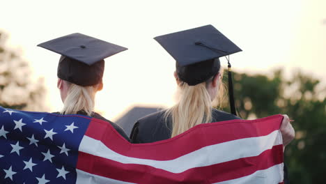 Zwei-Weibliche-Hochschulabsolventen-Mit-Amerika-Flagge-Auf-Den-Schultern-Rückansicht-Rear