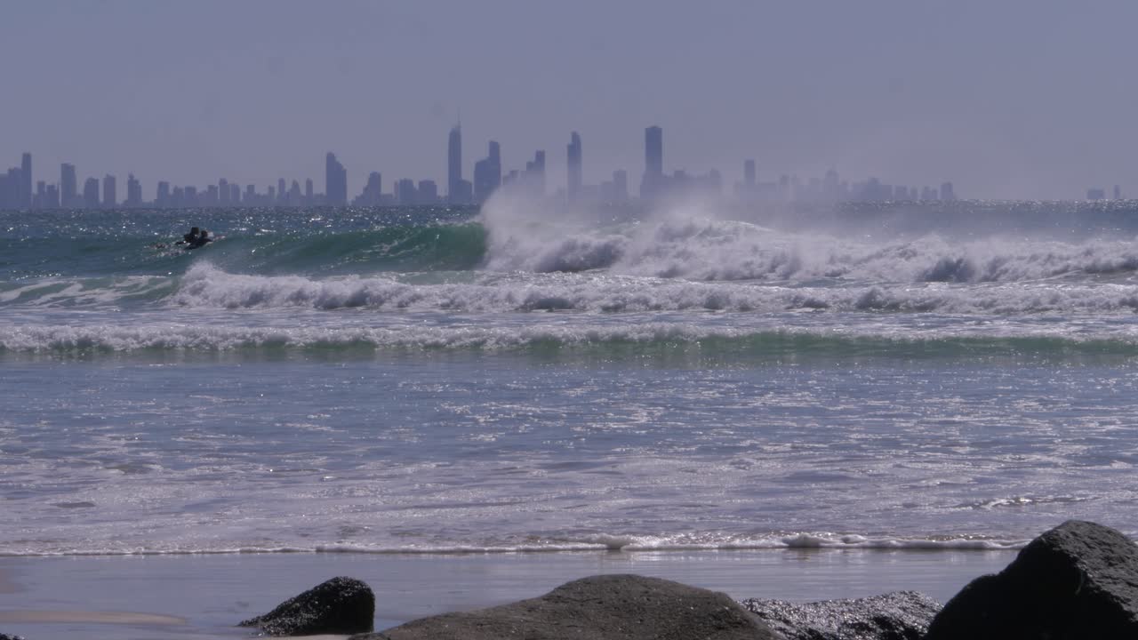Ocean Waves And Surfers With Gold Coast Skyline In Foggy Background. Rainbow Bay, QLD, Australia, wide shot