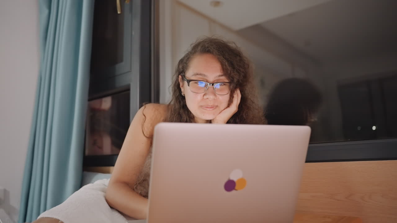 Noche en la habitación del hotel, sesión acogedora de navegación, mujer sonriente con portátil, viendo contenido y chateando, postura relajada en la cama cerca de la ventana, suave resplandor de la lámpara y reflejo de la cortina, ambiente de viajero casual con wifi y luz