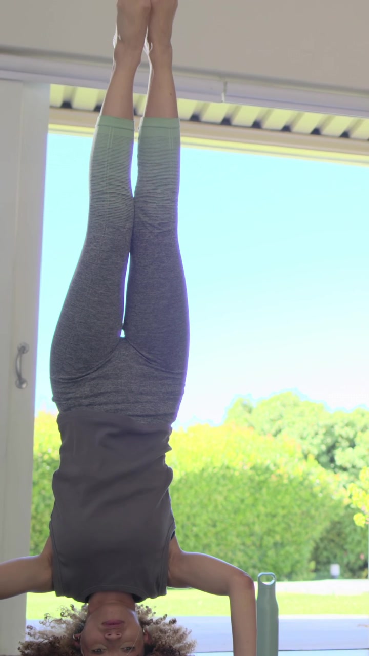 Vertical video: Leaving headstand, yogi in tank kneeling at glass doors, reaching bottle to sip