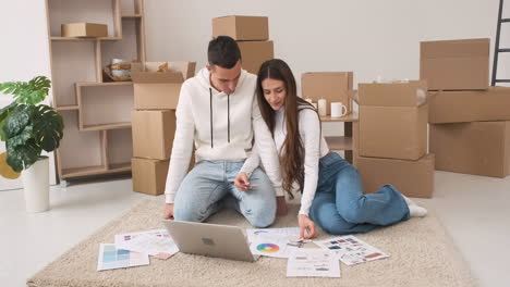 pareja joven en una casa nueva sentada en la alfombra con una laptop eligiendo colores para la decoración