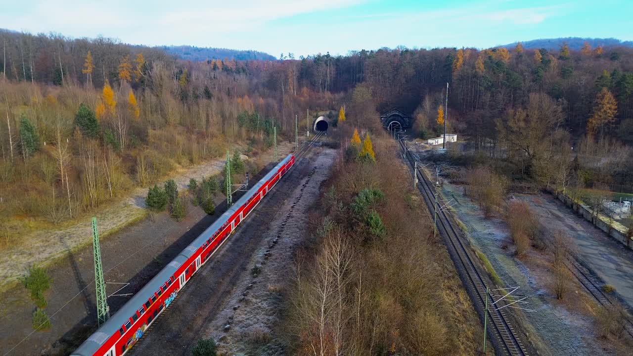 Red regional train approaching twin railway tunnels in bright winter landscape captured from aerial perspective