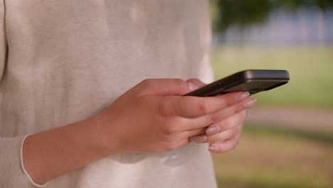 close-up of individual holding smartphone with polished nails, typing attentively while shadows fall gently on her light-colored clothing, blurred natural green background