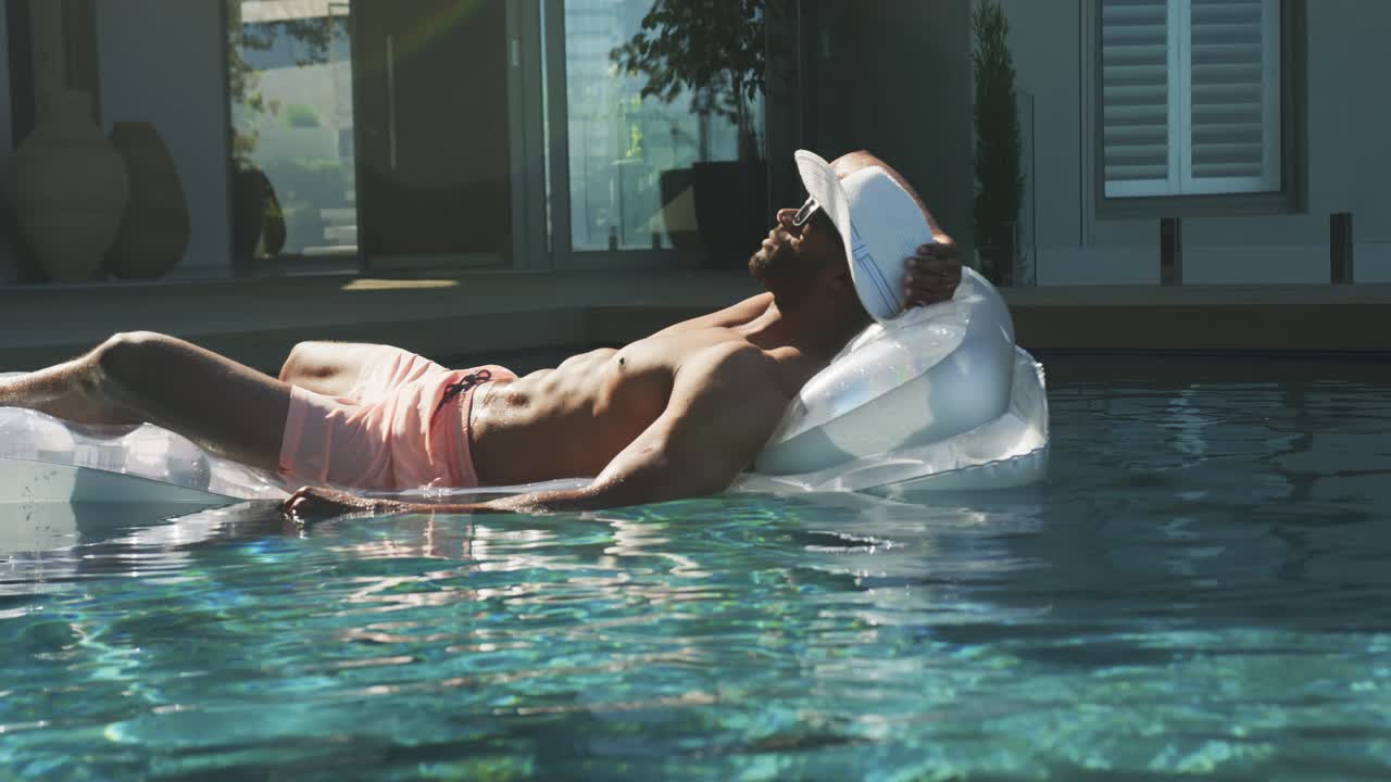 African American mature man relaxing on white lounger, drifting on pool ripples, steadying hat
