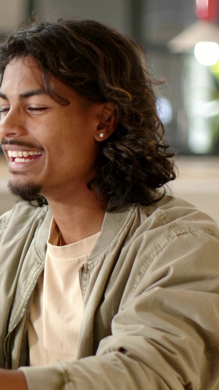 Vertical video: Smiling man in jacket typing on laptop at work table, glancing left with smartwatch