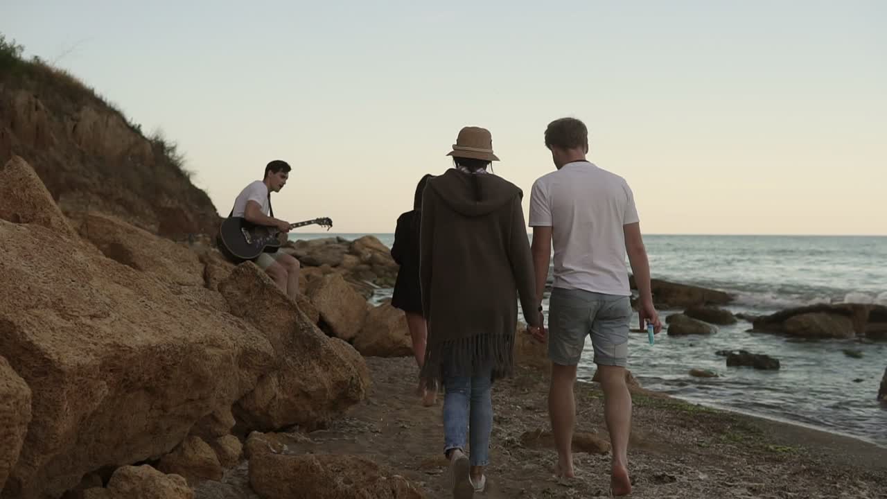 grupo de personas jóvenes y alegres caminando por la orilla del mar y sentados en las rocas por la noche, tocando la guitarra y cantando