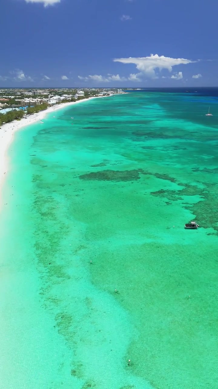 Drone verticale che sale e si sposta a destra sopra Seven Mile Beach, mostrando acqua turchese, sabbia bianca, la costa con resort e nuotatori