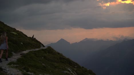 very cinematic footage of a blonde girl in a white dress walking on an exposed mountain path towards the rising sun until she disappears behind the mountain