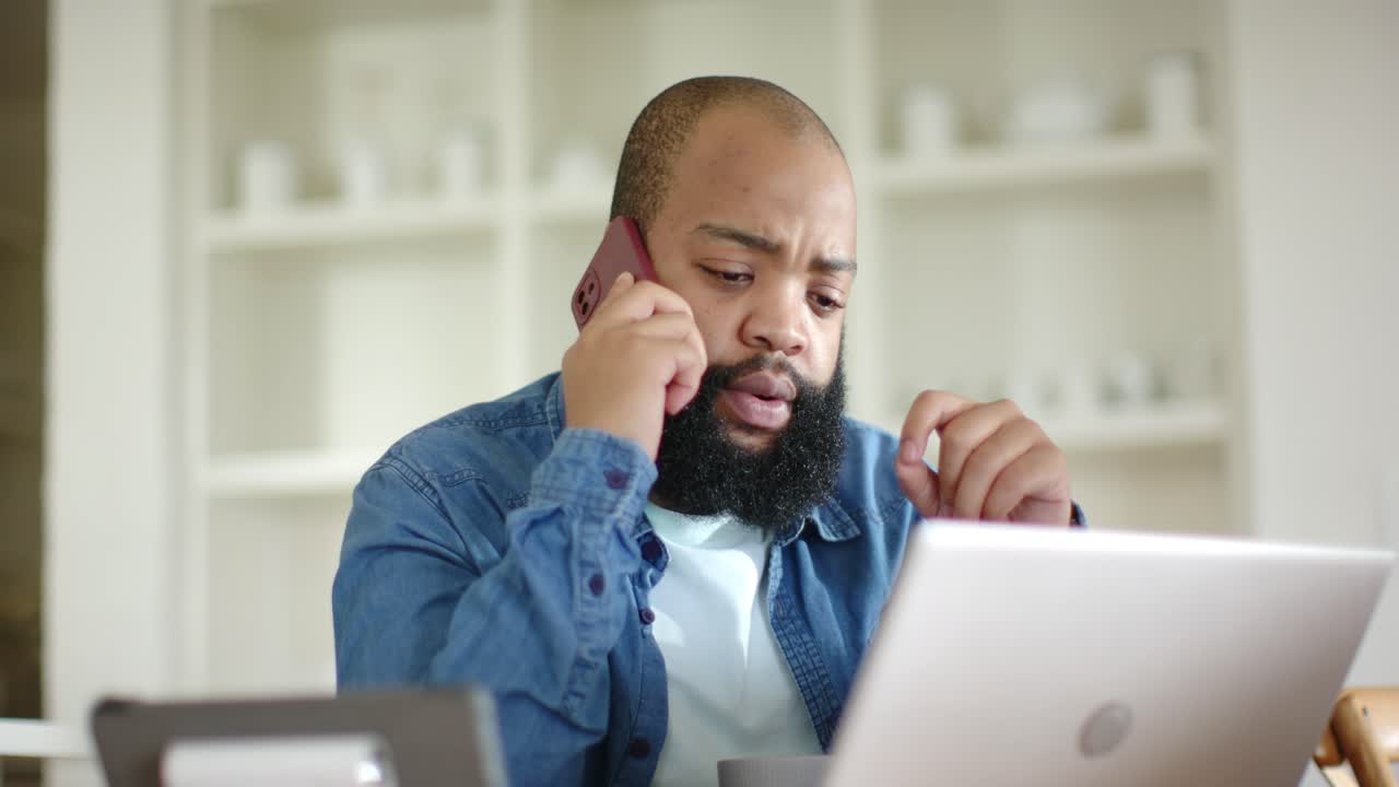 Vertical video: Lifting pink-case phone, man wearing denim shirt listening, checking laptop at home