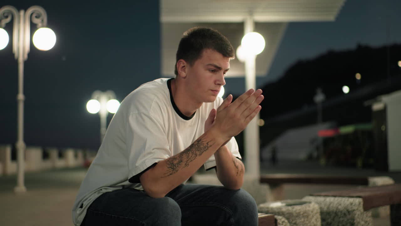 어두워지면서 문신한 손으로 벤치에 앉아 있는 반사적 인 남자, 밤에는 도시 불빛과 도시 구조물의 배경으로 사려 깊게보고 있습니다.