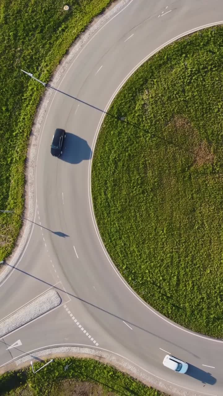 Vertical top down drone captures Katlakalns roundabout with a single white car tracing the curved road, its shadow stretching across the asphalt and grassy center under bright afternoon light