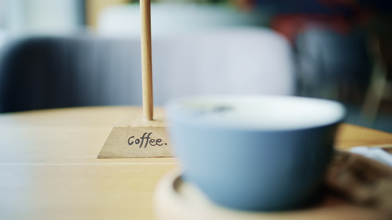 Piccolo cartello da tavolo in legno con la scritta "Caffè" posizionato su un tavolo da caffè con una tazza di caffè sfocata sullo sfondo.
