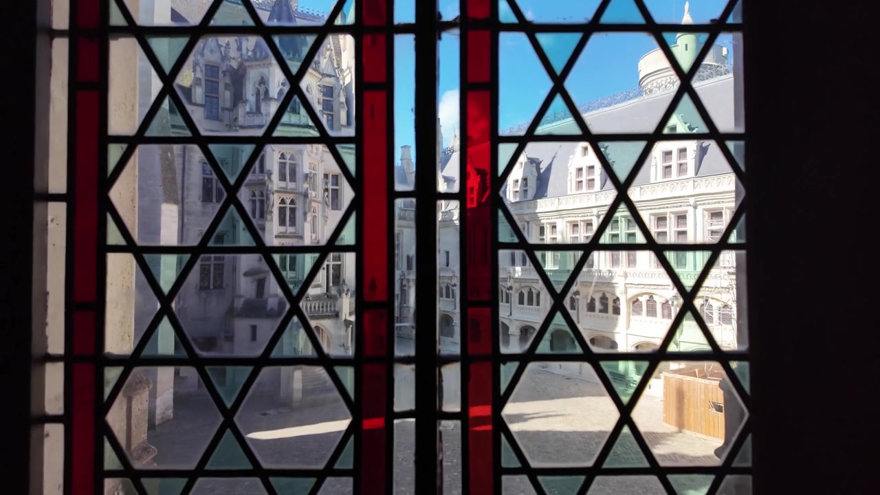 Vista attraverso una vetrata geometrica colorata al Château de Pierrefonds, che rivela facciate in pietra decorate, abbaini, archi e il cortile interno