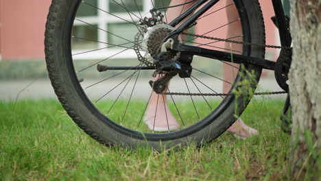 close-up of bicycle wheel and pedal with person wearing pink sneaker removing stand in lush green park with blurred background of grass, trees, and building