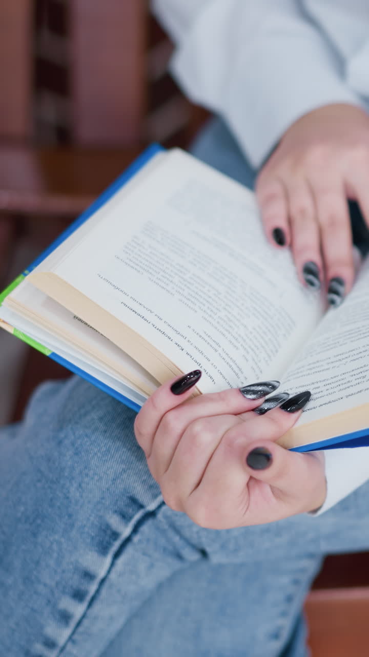primer plano de un estudiante con uñas pintadas sosteniendo un libro en la pierna, preparándose para volcar una página, con camisa blanca y vaqueros, sentado en un banco de madera con una vista parcial de la bolsa de compras rosa en el fondo