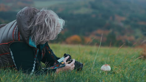 female photographer with grey hair taking pictures of parasol mushroom in the green grass in autumn nature during a cold windy day surrounded by colourful trees in slow motion