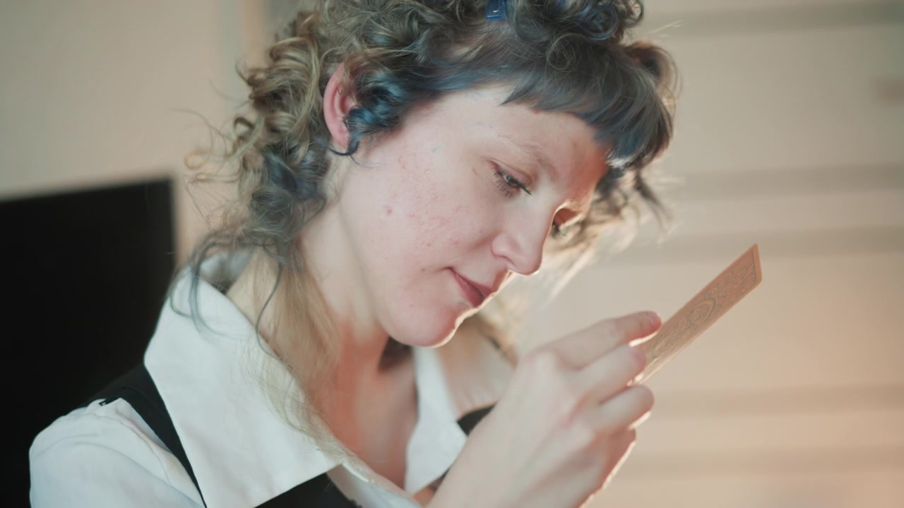 Primo piano intimo di una donna che studia una carta in luce soffusa, capelli ricci con striature blu, mazzo vintage tenuto tra dita attente, espressione concentrata che suggerisce una lettura privata della fortuna e un tranquillo rituale di preveggenza.