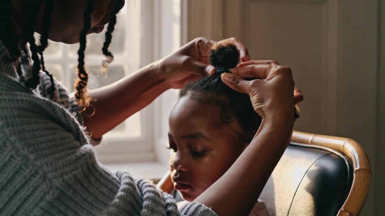 Gathering caregiver twisting girl's hair into top bun at home, securing tie, child in pink top