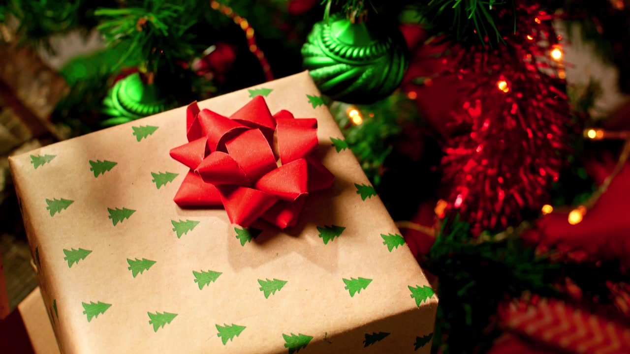 Tomada em tilt suave de um presente de Natal embrulhado sob uma árvore decorada com luzes brilhantes, enfeites vermelhos e verdes, guirlandas e uma atmosfera festiva e aconchegante.