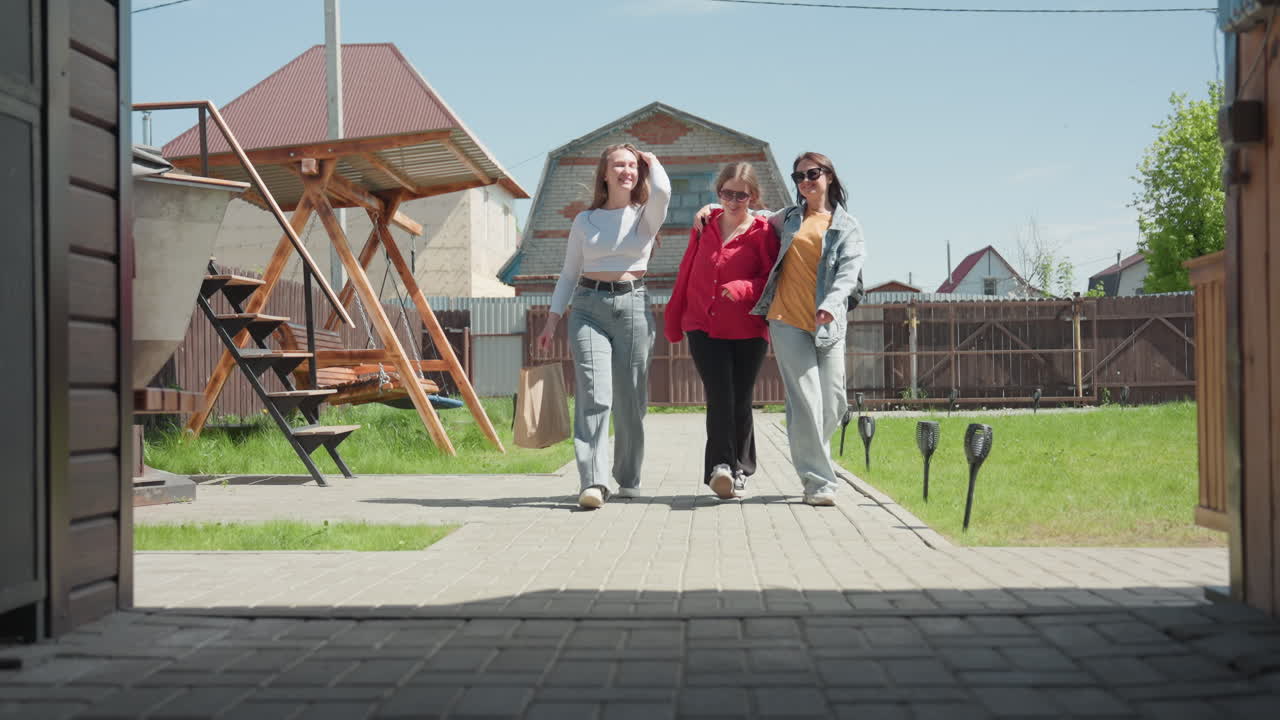 Girls walk into recreational center along paved walkway, smiling and chatting while one holds arm of another, passing wooden swing under metal roof in sunny suburban yard with green grass and fence