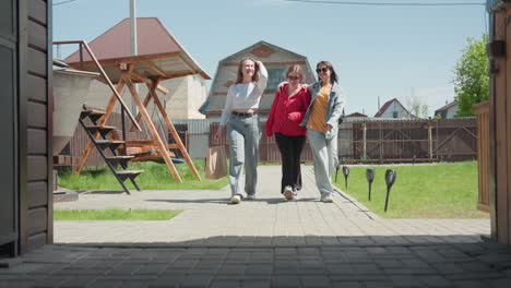 girls walk into recreational center along paved walkway, smiling and chatting while one holds arm of another, passing wooden swing under metal roof in sunny suburban yard with green grass and fence