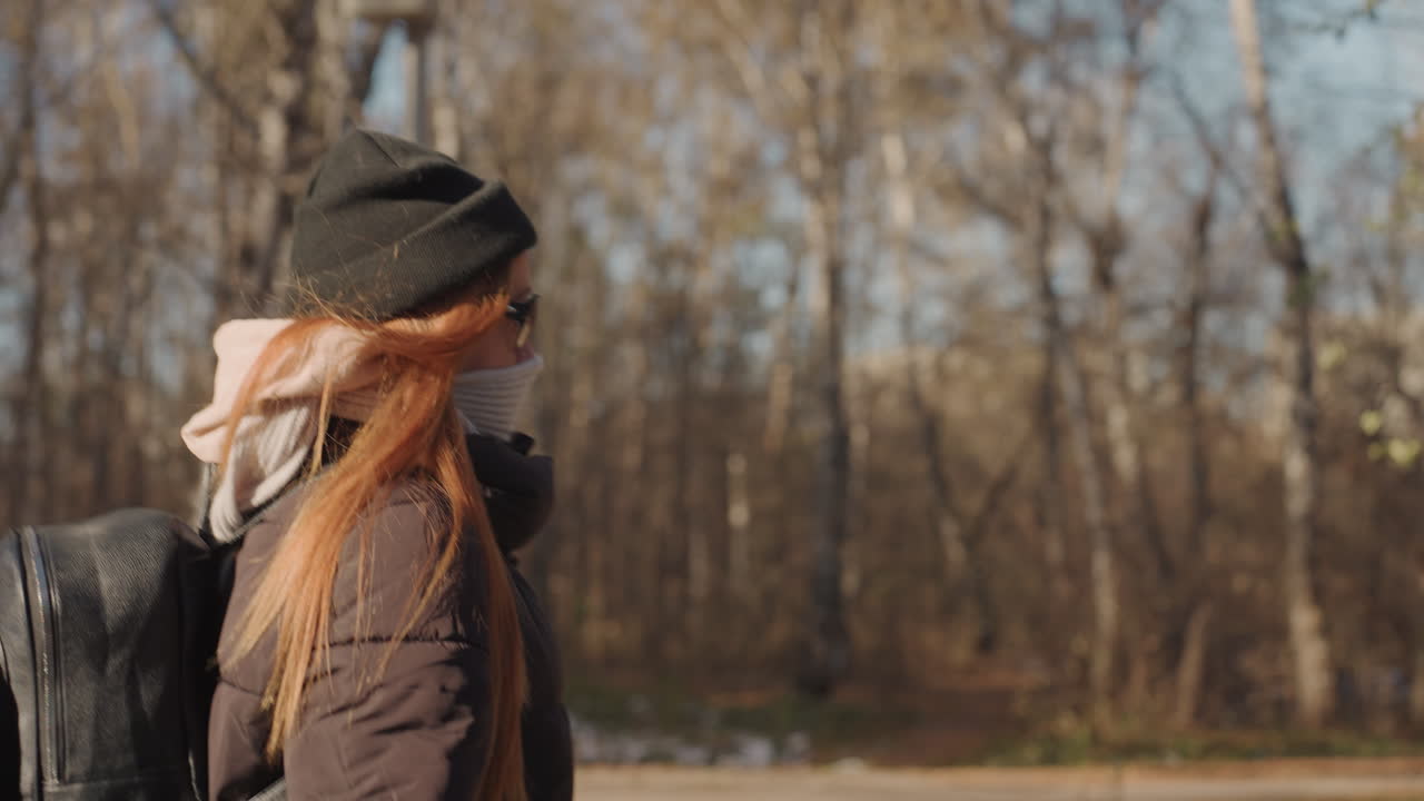 Caucasian woman frontal closeup walking through treelined avenue wearing sunglasses, mask and beanie, long red hair flowing and carrying a case backpack, assertive commuter stride with empty path