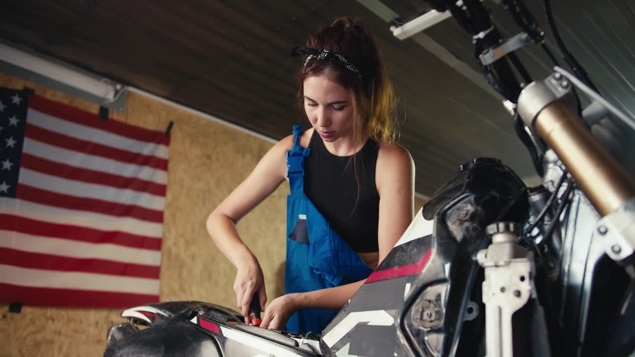 una chica mecánica morena con una camiseta negra y un mono azul repara una motocicleta en su taller contra el telón de fondo de la bandera de los estados unidos. trabajando como mecánica, chica reparando una motocicleta