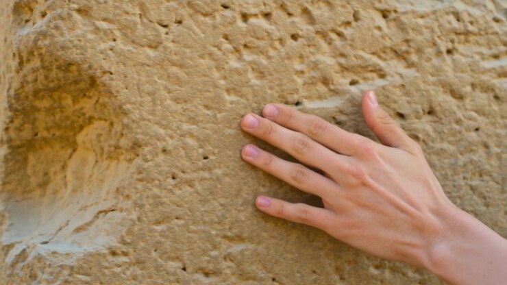 Approaching bare right hand pressing and sliding across tan stone block at ruins, feeling erosion