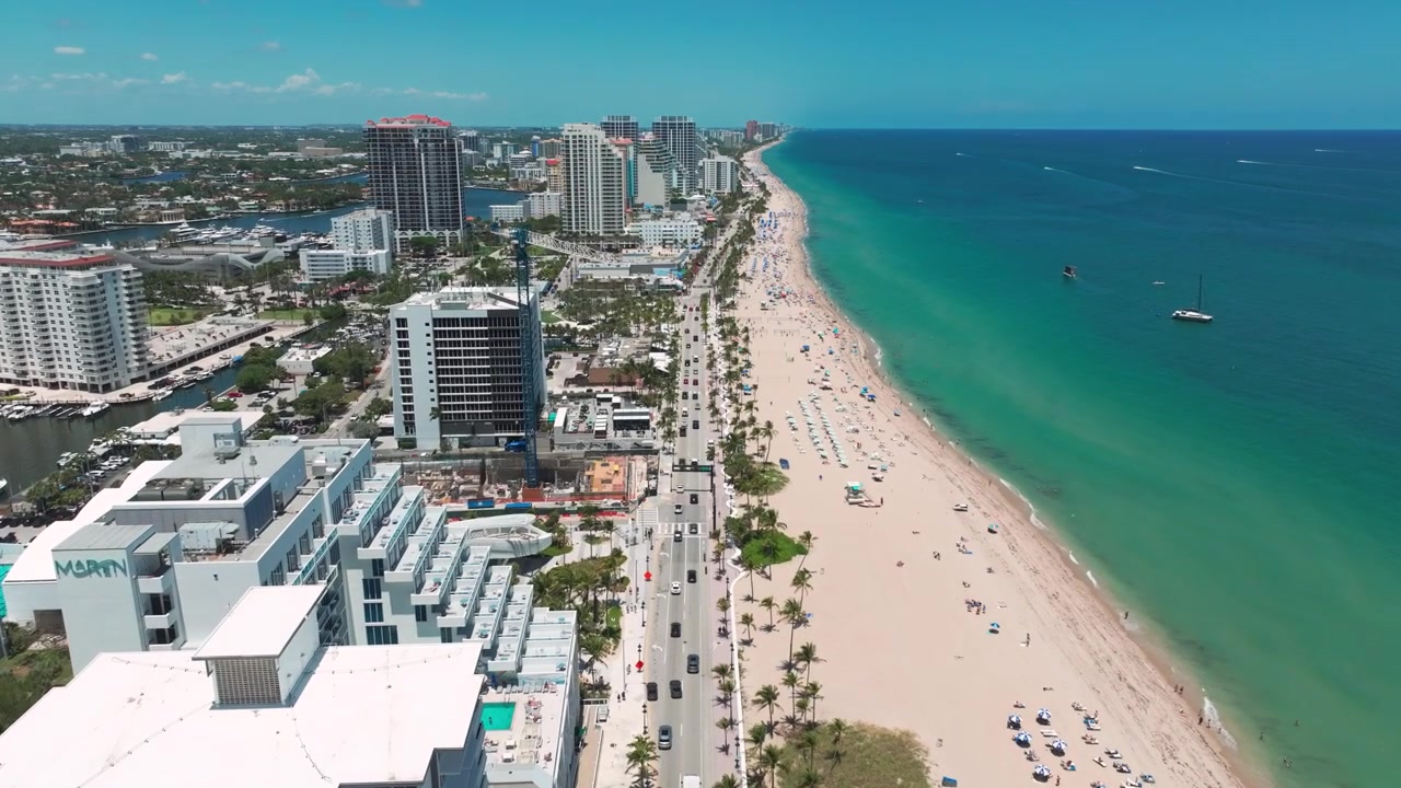 Perspectiva soleada de dron de la playa de Fort Lauderdale, capturando hileras de palmeras, sombrillas de playa, complejos turísticos frente al mar y un flujo constante de tráfico a lo largo de la A1A