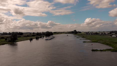 Aerial-showing-large-cargo-vessel-ship-on-river-IJssel-with-Dutch-flag-outside-Hanseatic-city-Zutphen,-The-Netherlands,-against-a-blue-sky-with-clouds