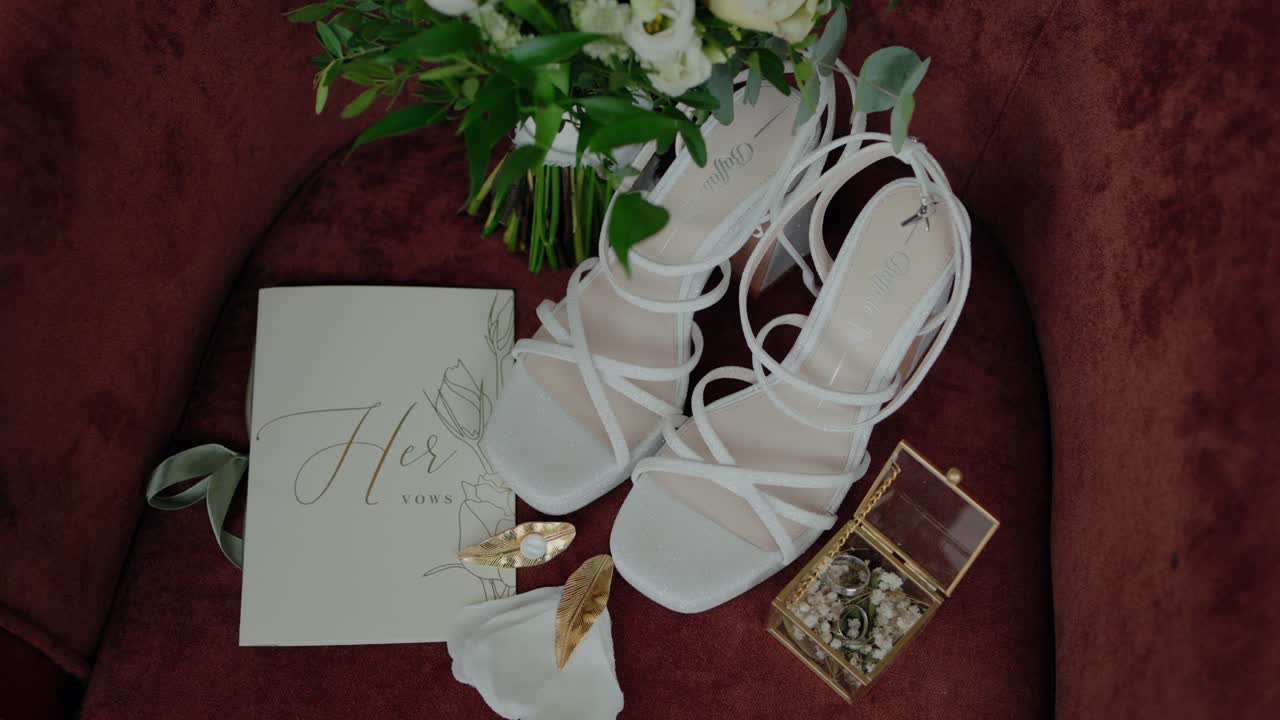 Elegant bridal essentials beautifully arranged, showcasing white heels, a bouquet, wedding vow book, and a jewelry box on a luxurious velvet chair