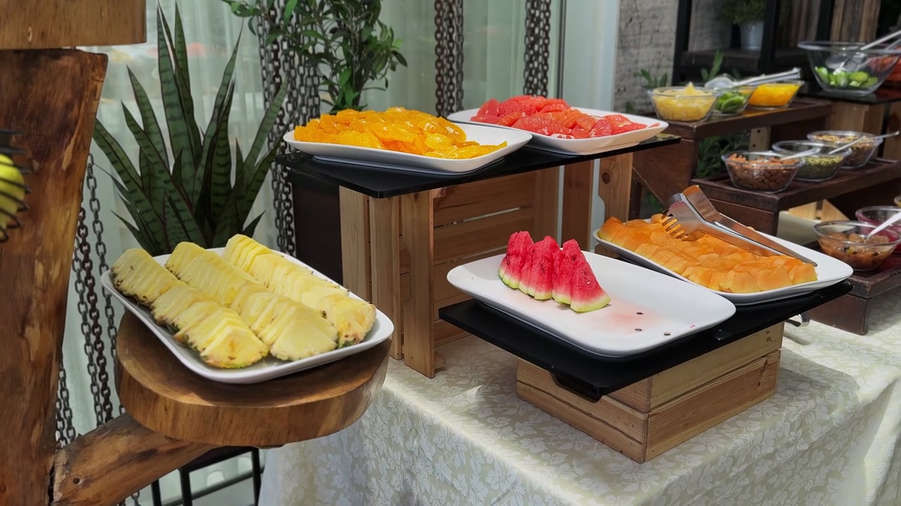 Vista luminosa diurna di un buffet di frutta fresca a fette su vassoi bianchi e alzate in legno a più livelli. L'esposizione colorata e salutare per la colazione presenta ananas succoso, anguria e arance in un hotel.