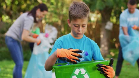 Smiling-caucasian-son-outdoors-holding-recycling-crate,-collecting-plastic-waste-with-parents