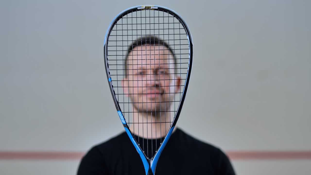 Man lifts squash racket in front of face, smiling in creative sports portrait