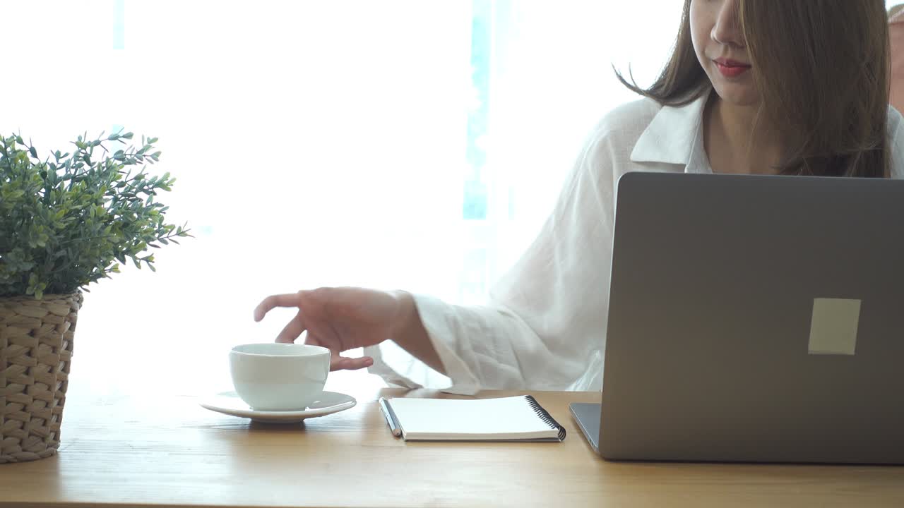 自宅のリビングルームで温かいコーヒーを楽しみながらノートパソコンで作業する美しい若い笑顔の女性。家での時間を満喫。自宅オフィスで働くアジア人ビジネスウーマン。