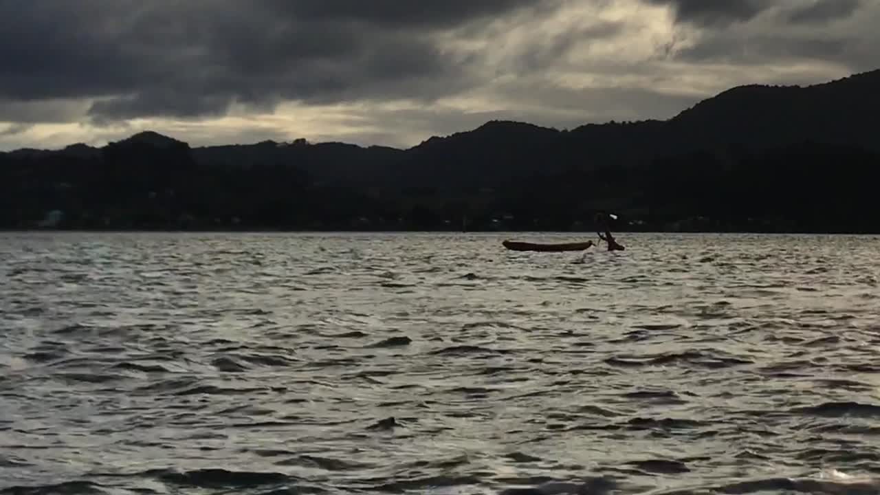 실루엣의 새가 가로질러 날아가는 동안 카약은 산을 배경으로 석양에 졸졸 흐르는 호수 표면에 부드럽게 떠 있습니다.