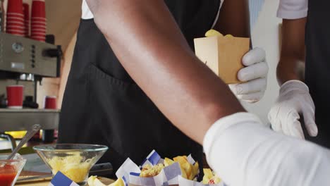 sección media de un hombre afroamericano con un delantal poniendo papas fritas en una caja en el camión de comida