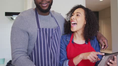 Glückliches-Afroamerikanisches-Paar-Trägt-Schürzen,-Benutzt-Tablet-In-Der-Küche-Und-Redet