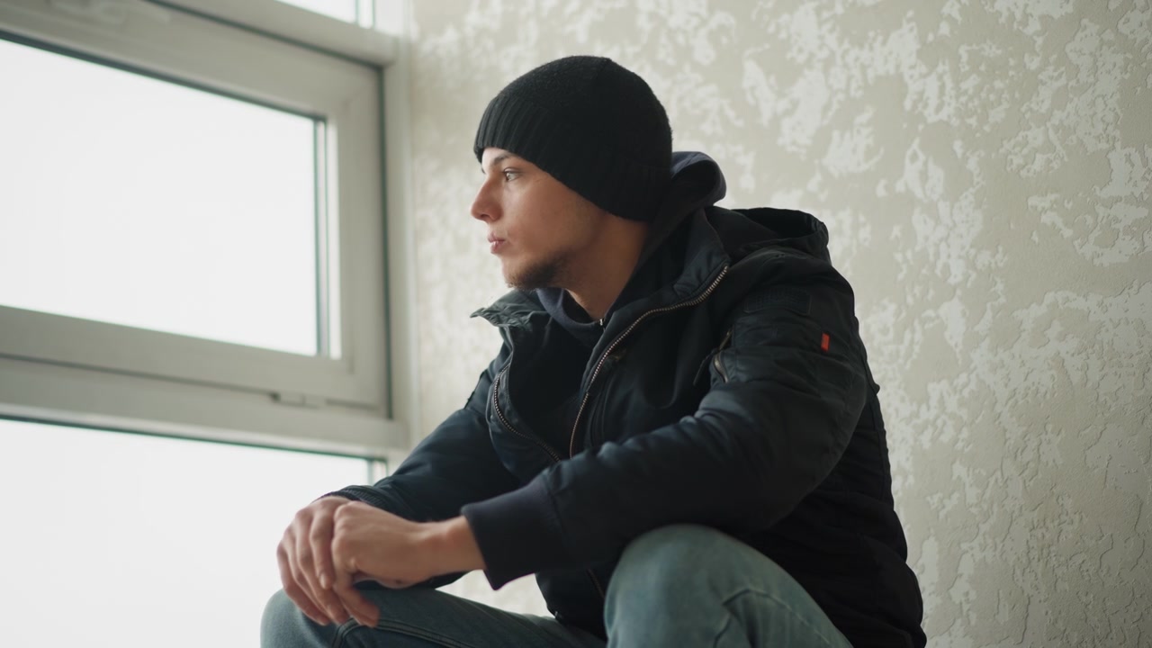 Close up of young man sitting by window wearing dark clothing and looking out. scene is indoors with soft natural light from outside featuring textured walls and minimal camera movement