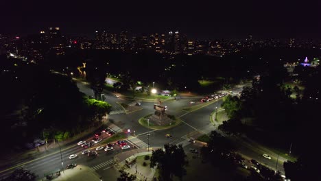 Aerial-view-establishing-at-night-of-the-Urquiza-Monument-in-the-middle-of-Palermo's-parks,-low-vehicular-traffic