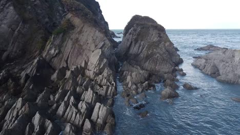 Aerial-drone-flying-over-water,-out-to-sea,-through-large-rock-formations-under-a-cliff---Lee-Bay,-Beach,-Ilfracombe,-Devon,-England