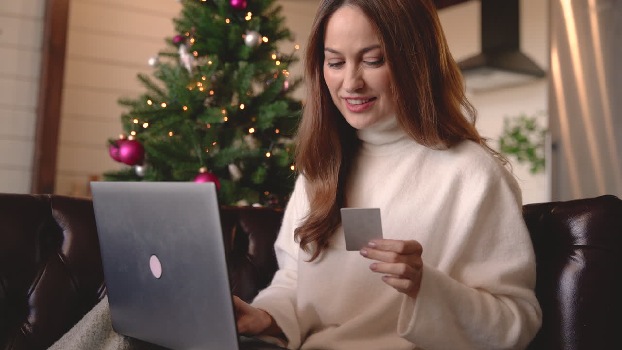 mujer feliz comprando en línea usando tarjeta de crédito mientras se sienta en el sofá cerca de un árbol de navidad en la sala de estar en casa