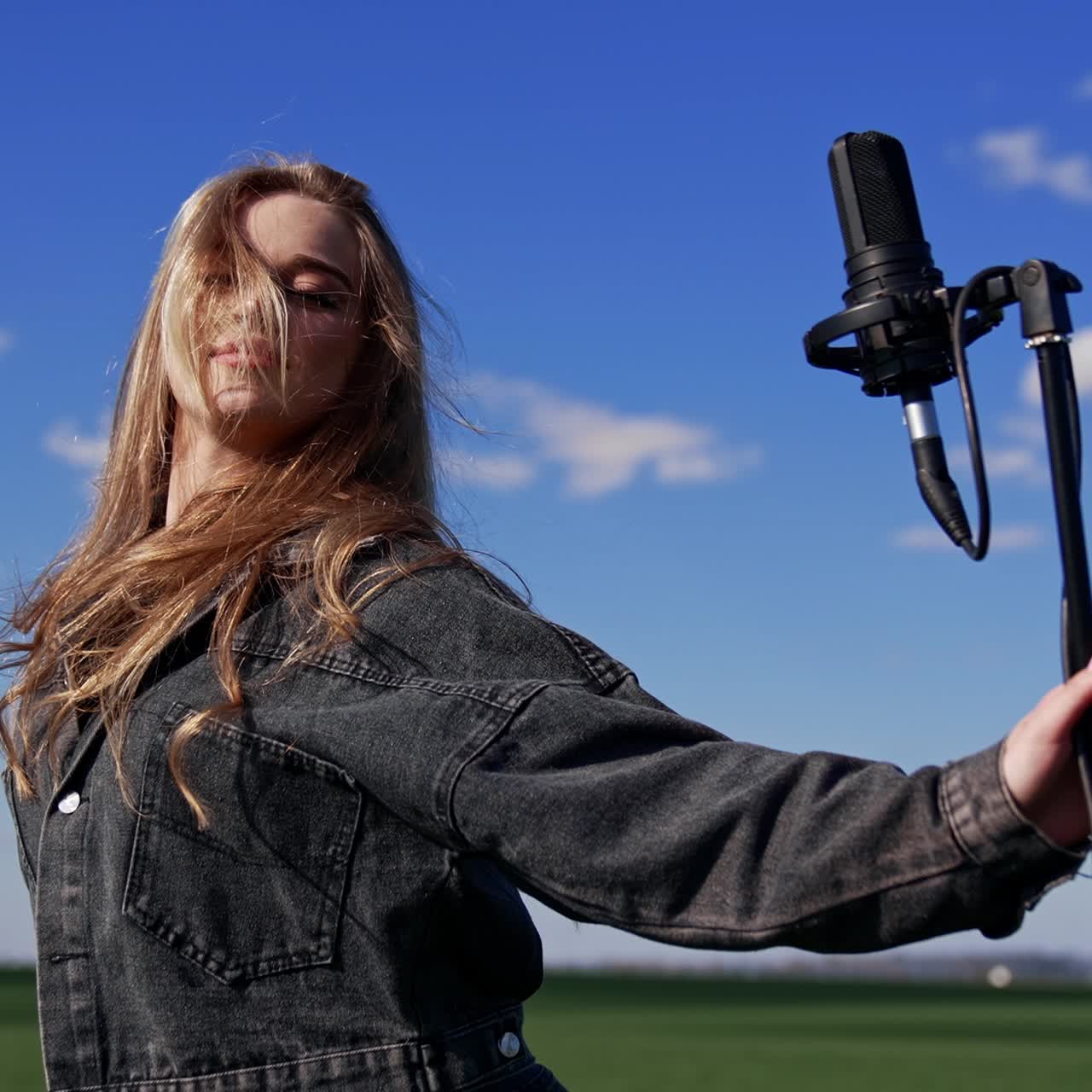 Attractive girl with microphone on field. Sexy young model with long hair in denim suit standing near the microphone and dancing looking at camera outdoors