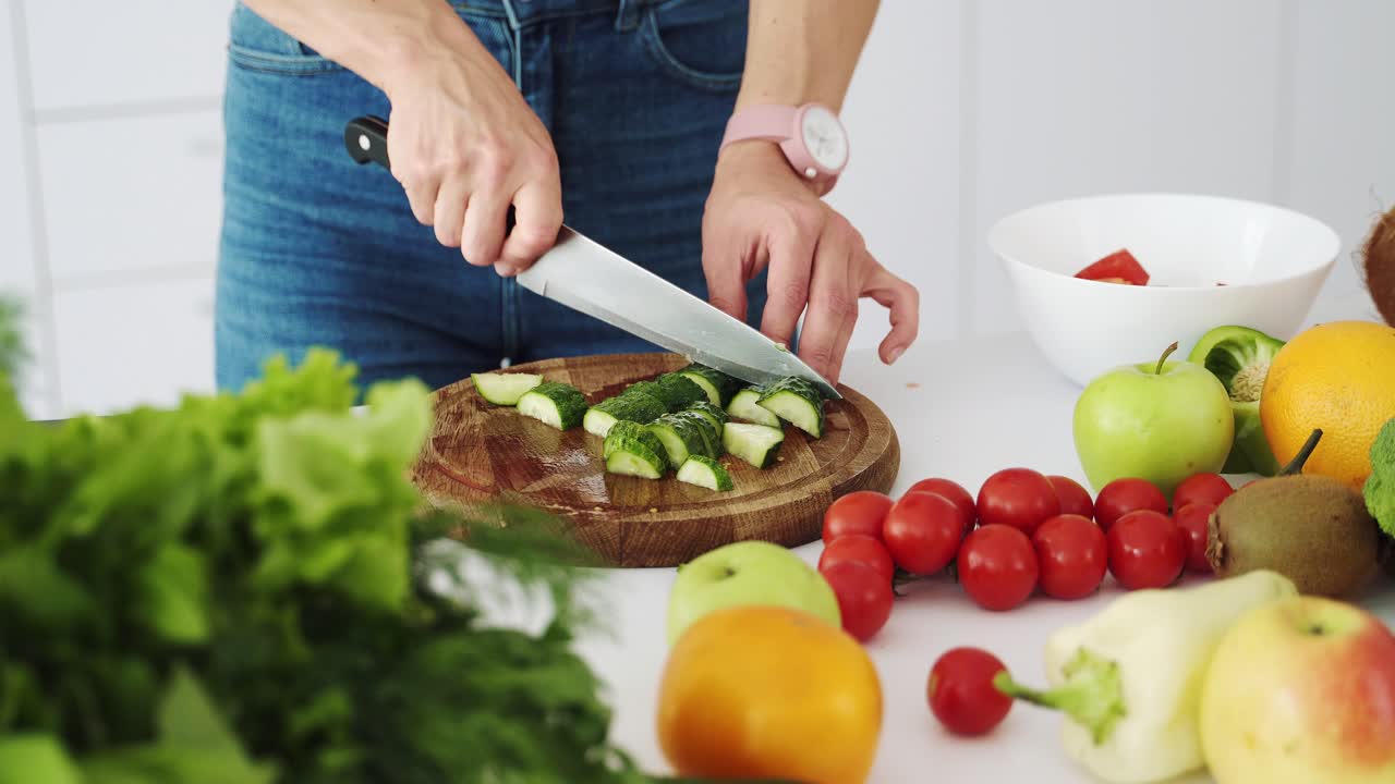 Female cook cuts a fresh green cucumber with a knife on a wooden cutting board. Table with vegetables and fruits for making salad