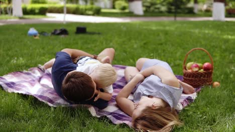 Loving-young-family-with-little-son-have-a-picnic-outdoors.-Green-park,-picnic-blanket-with-basket-full-of-fruits,-Father-and-mother-falling-down-on-a-grass.-Father-holding-boy-in-his-hands.-Backside-view