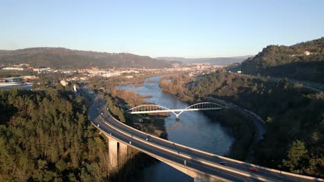 Aerial-shot-over-a-bridge-and-a-freeway-in-the-River-Miño-in-Spain,-great-vegetation,-horizon-with-mountains-and-blue-sky