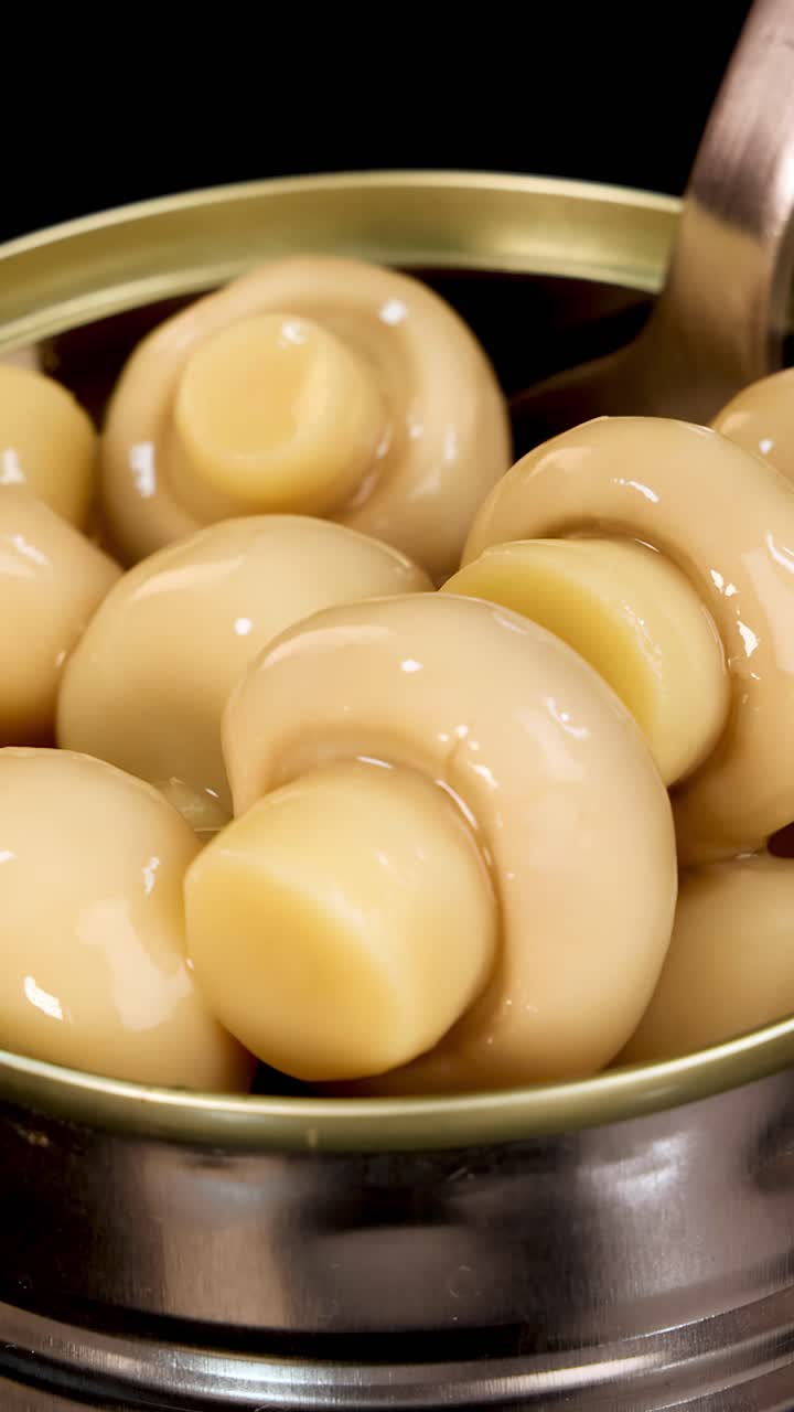 Un cucchiaio di metallo solleva funghi champignon interi da una lattina aperta sotto un'illuminazione brillante e uniforme, evidenziando la texture lucida e l'ambiente del cibo conservato.